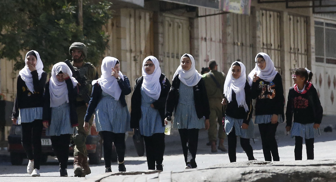Photo of an Israeli soldier walking behind young women in school uniforms, some wearing hijabs.