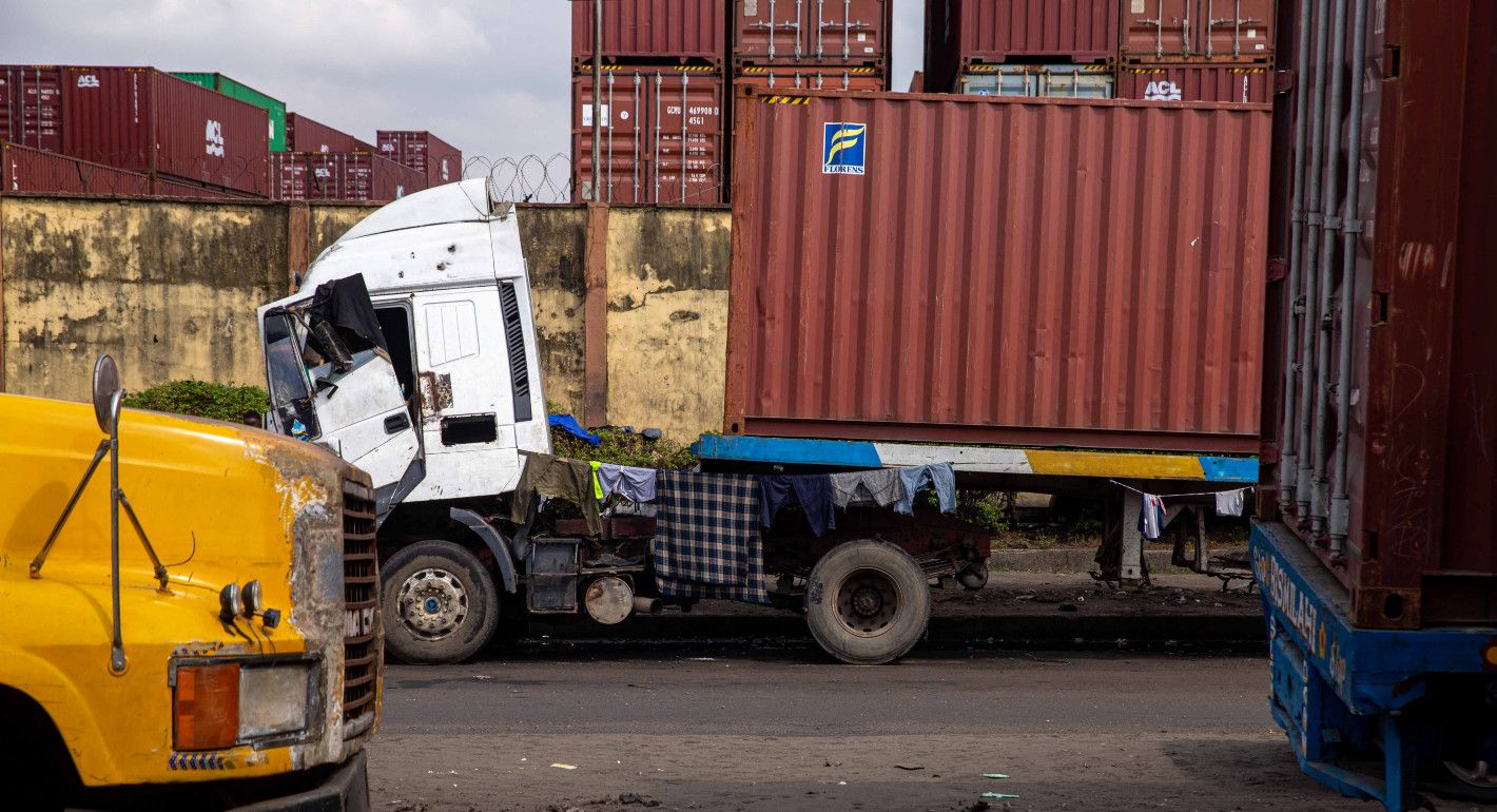 A large transport truck with a shipping container sits in the foreground. Stacks of shipping containers are visible in the background.