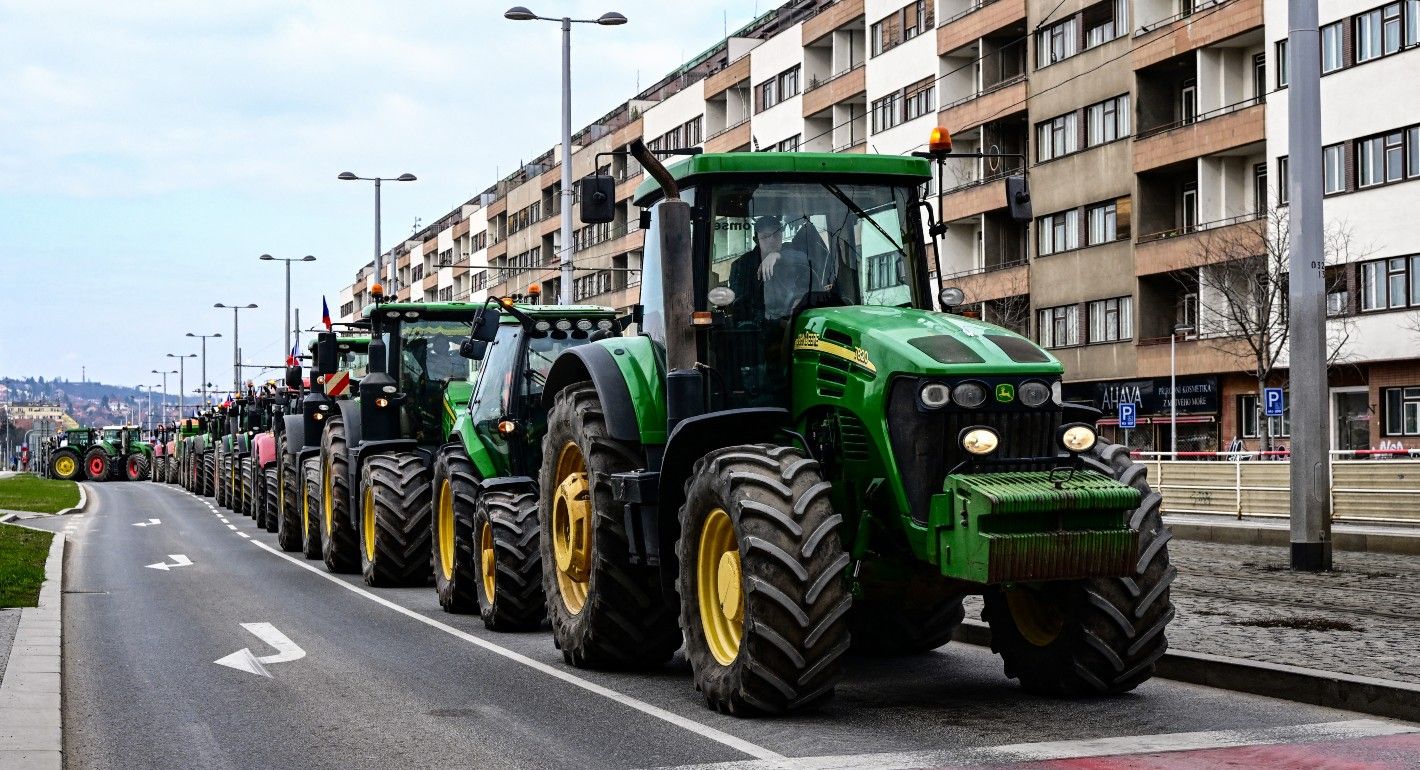 A row of large green trackors drive on a road made for cars. There is a block of apartment buildings in the background
