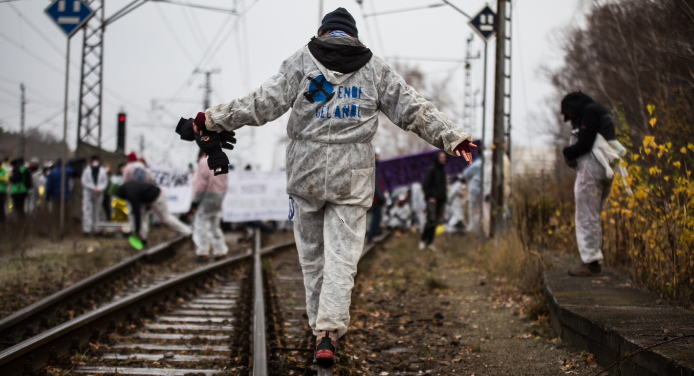 A person in a white jumpsuit with the words and logo of "Ende Gelände" balances walking along a railroad track with their back to the camera