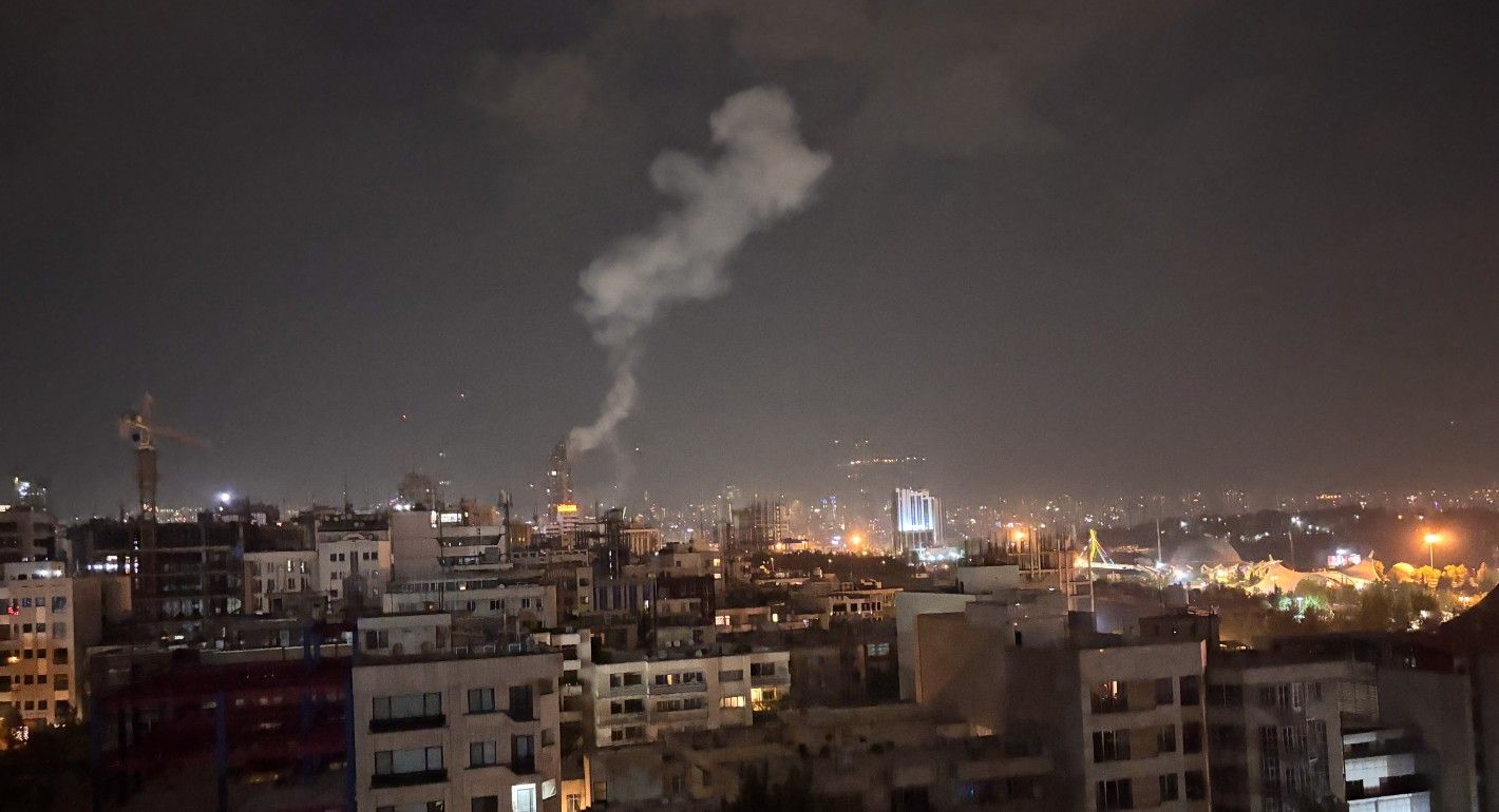 A plume of light gray smoke rises above Tehran's skyline against a dark gray morning sky