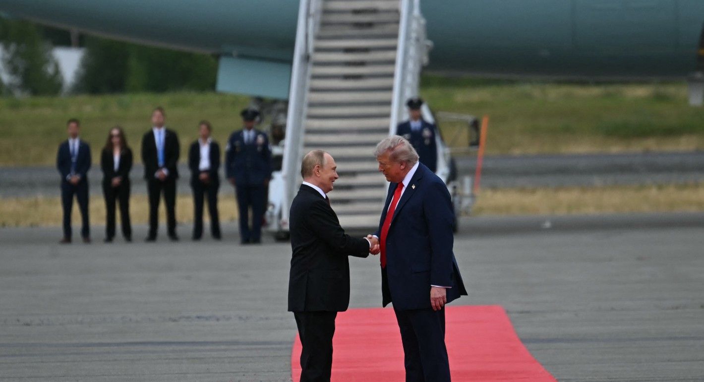 Trump and Putin stand on a red carpet in front of the loading stairs to an airplane. They shake hands.
