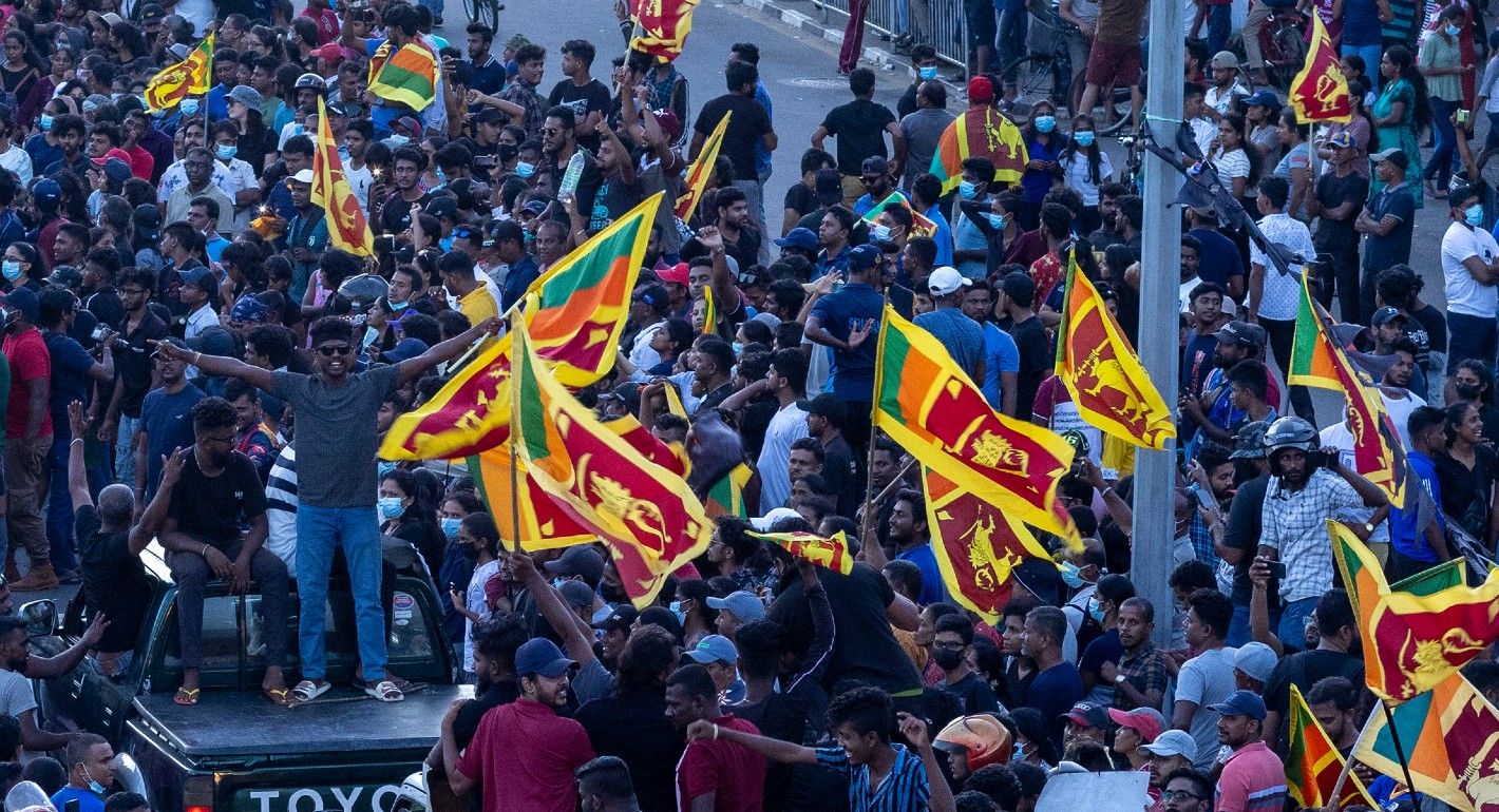 A crowd of Sri Lankan protesters hold Sri Lankan flags. One man stands atop a pickup truck on the left side of the frame.