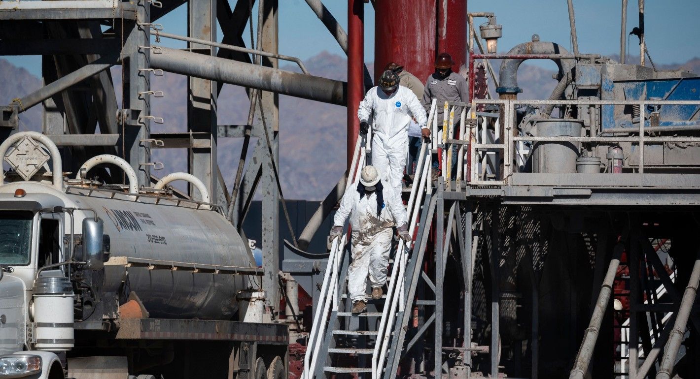Several people in white work suits and white hard hats climb down metal stairs on a drilling rig.