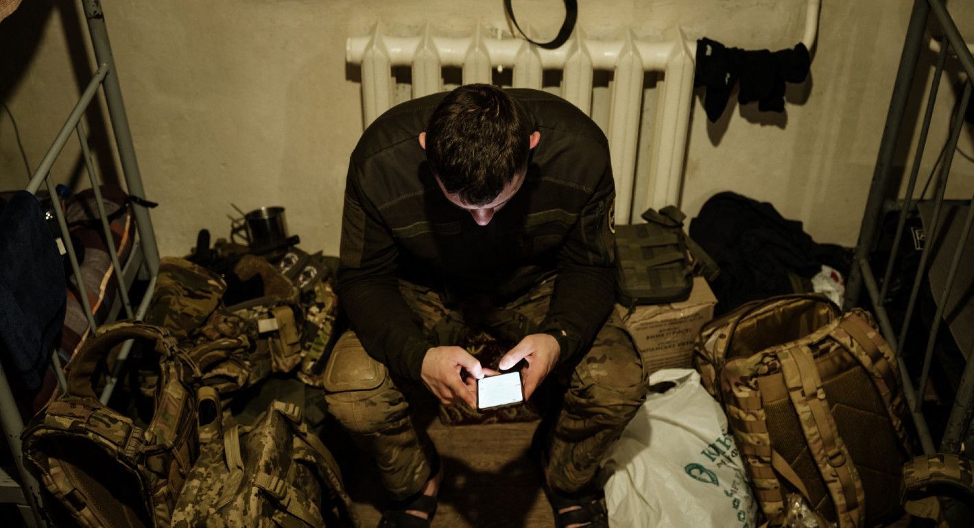 Photograph of a soldier, surrounded by gear, looking down at a smartphone.