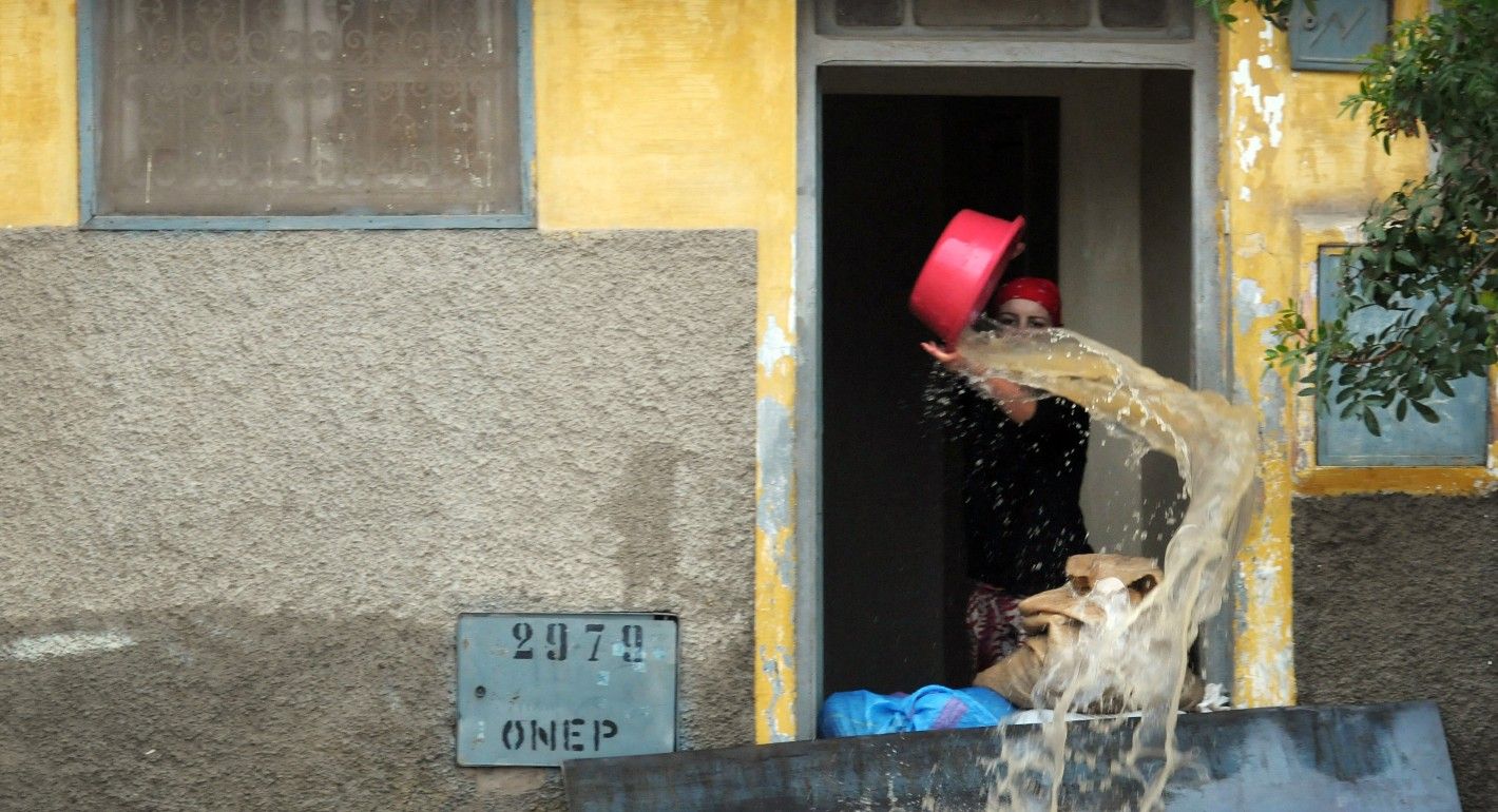 A woman holds a red bucket of water upside down, baling water from a yellow building into a flooded street